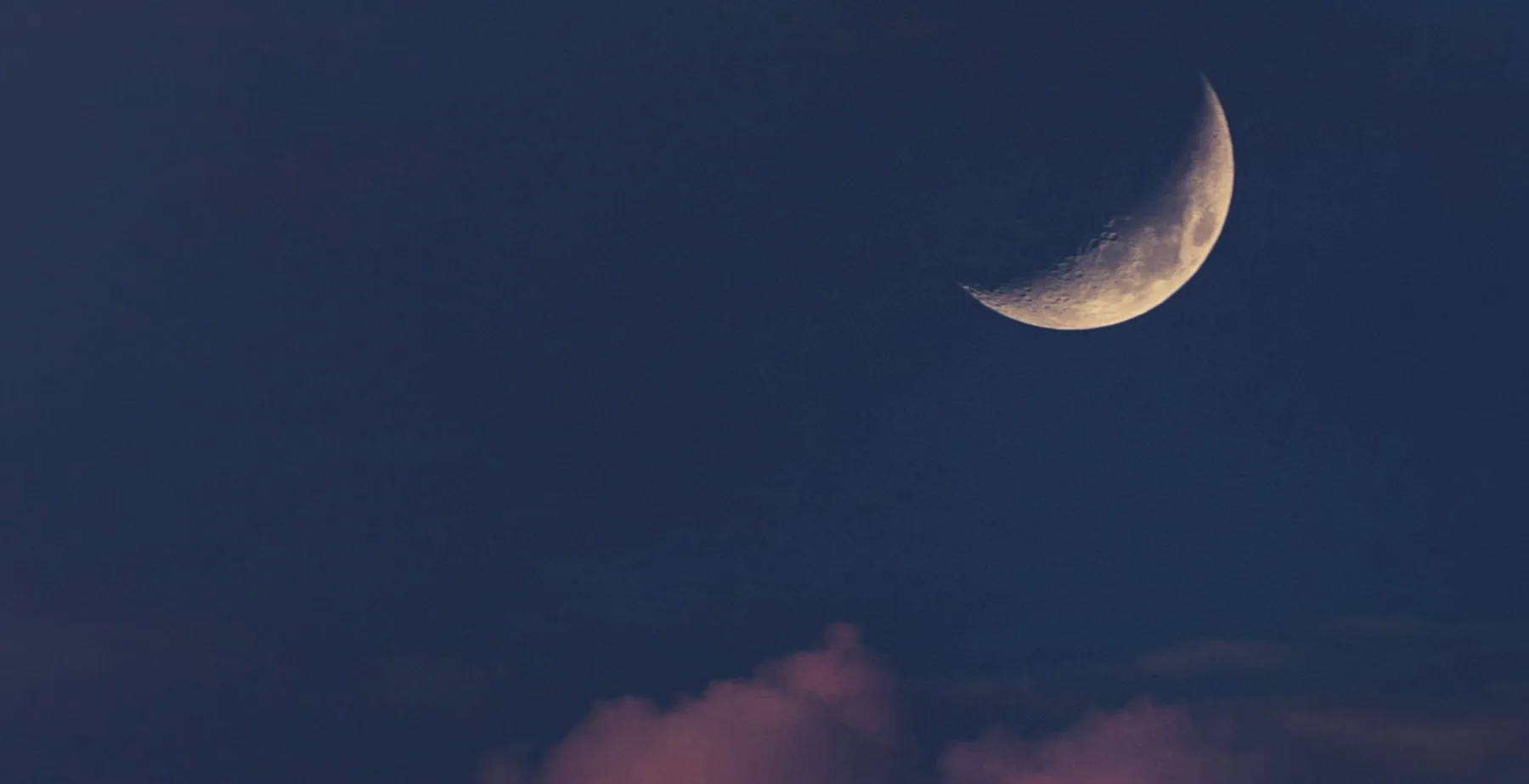 A crescent moon in a dark blue sky above a bank of purple cloud